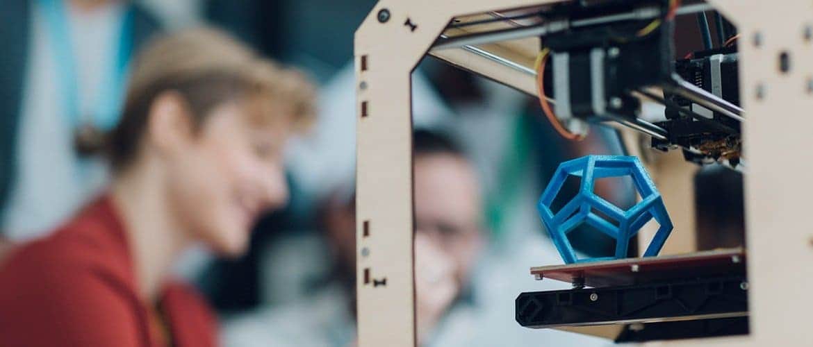 Imagem mostra uma impressora 3D em primeiro plano e pessoa sorrindo no segundo plano.