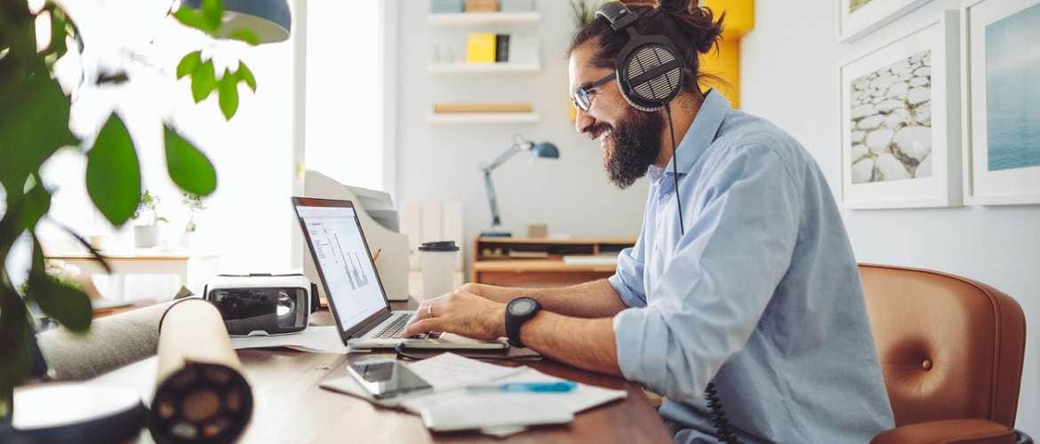 Homem realiza trabalho em casa, a partir de seu laptop, enquanto utiliza um fone de ouvido