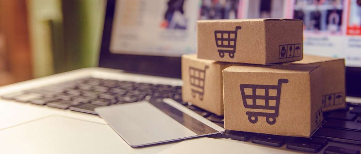 Imagem conceito de um e-commerce: um cartão de crédito está apoiado sobre o teclado de um laptop e, próximo a ele, existem três caixinhas com o ícone de um carrinho de supermercado sobre a superfície