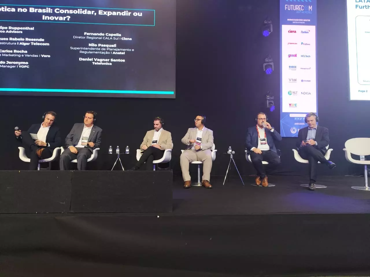 Painel de discussão sobre a expansão da fibra óptica no Brasil no Futurecom 2025, com vários especialistas e painel de monitoramento exibido ao fundo.