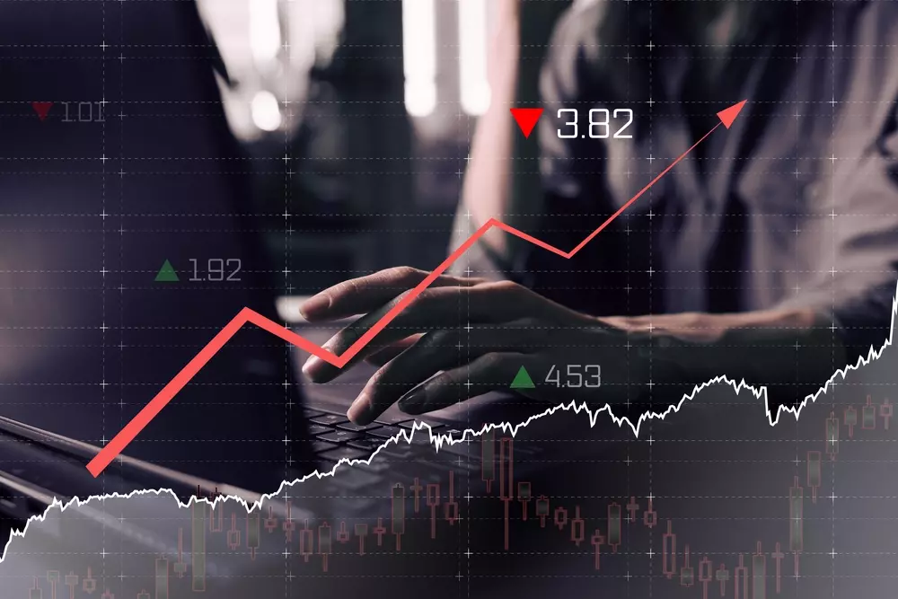 Pessoa digitando em um laptop enquanto gráficos financeiros e linhas de mercado aparecem sobrepostos na tela, incluindo setas vermelhas e verdes indicando variações de valores e tendência de alta.