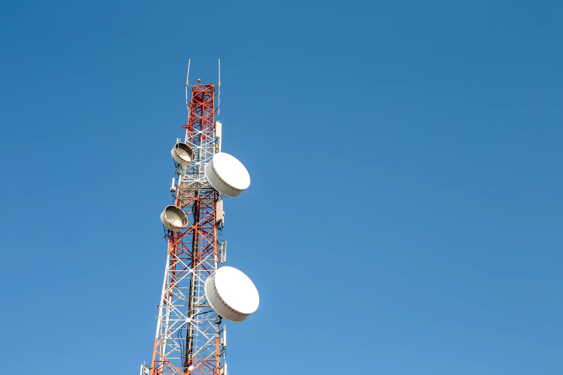 Torre de telecomunicações com antenas de transmissão no topo, estrutura metálica pintada em azul e vermelho contra céu azul, tecnologia de comunicação sem fio