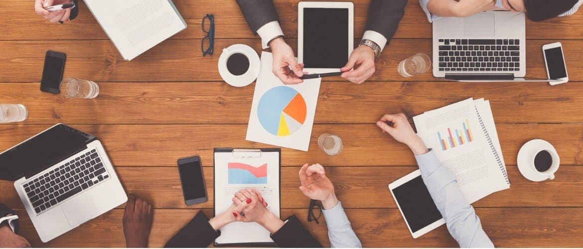 Imagem de uma reunião em ambiente corporativo com as pessoas utilizando laptops, tablets, celulares e gráficos impressos em papel