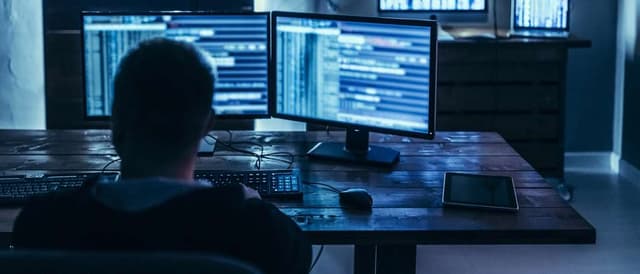 Imagem de uma pessoa observando duas telas de computador em um cenário escuro