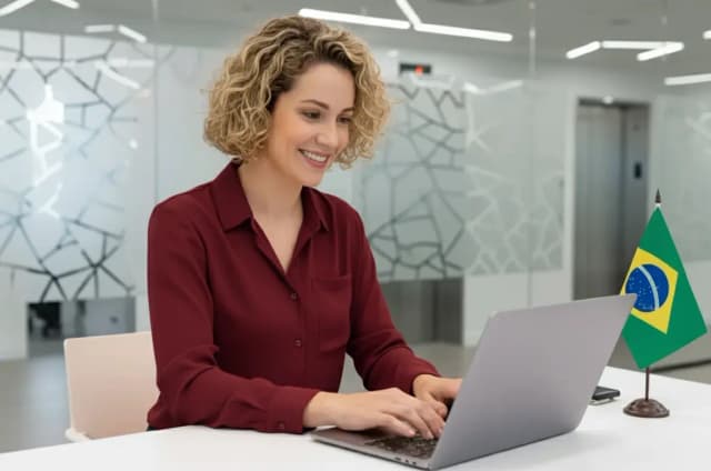 Mulher sorridente usando laptop em ambiente moderno com bandeira do Brasil ao lado, simbolizando profissionalismo e conexão com o país.