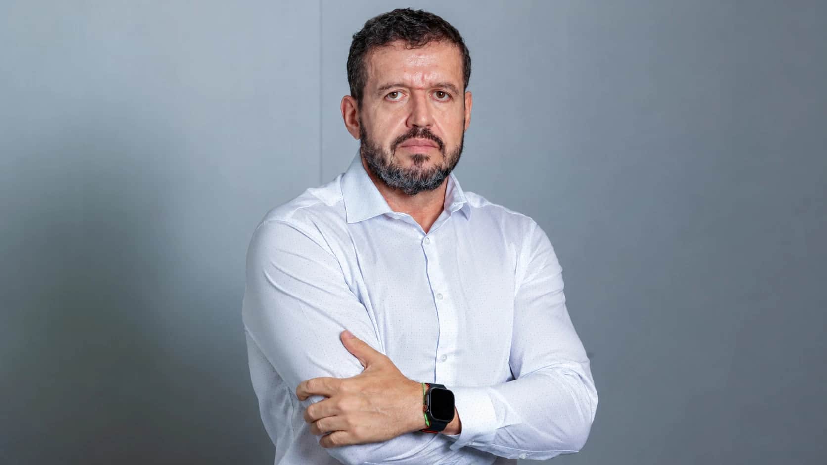 Foto do executivo Vitor com barba e cabelo escuro, usando camisa social branca, posando com o braço cruzado e relógio no pulso diante de fundo cinza, relacionada à Gerdau.