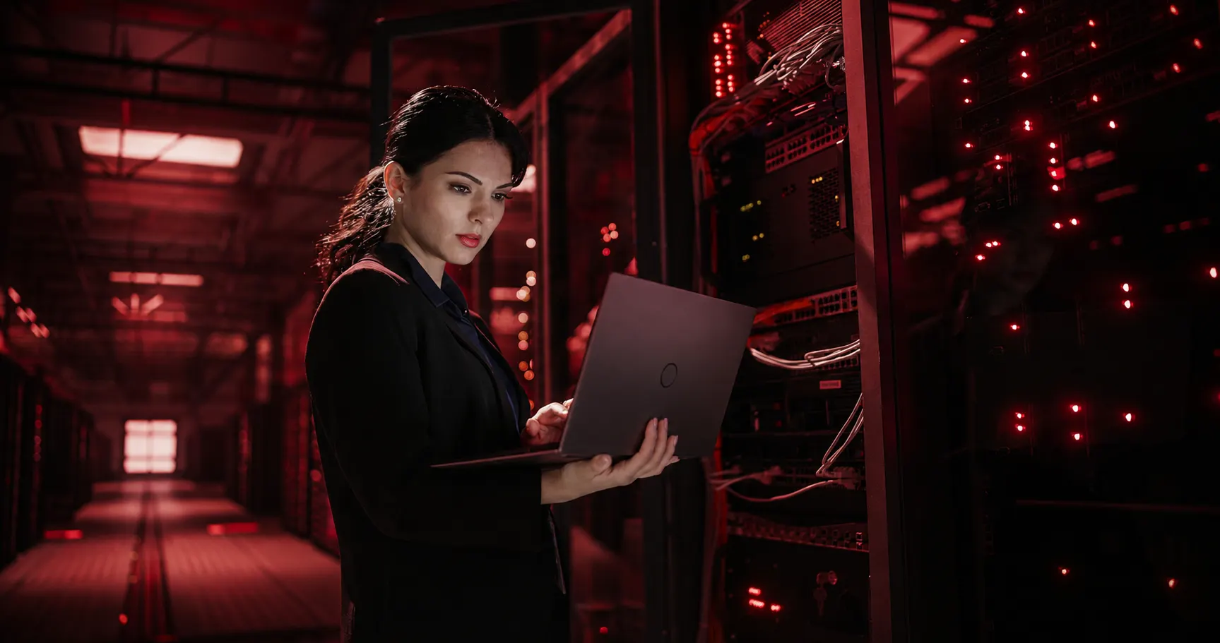Profissional de TI em data center observa um notebook enquanto ao fundo servidores e racks com luzes vermelhas indicam infraestrutura digital e computação em nuvem