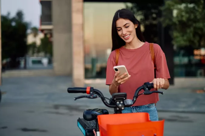 Jovem usando smartphone ao lado de bicicleta elétrica em ambiente urbano moderno, destacando a conectividade e inovação tecnológica nas cidades inteligentes habilitadas por 5G.