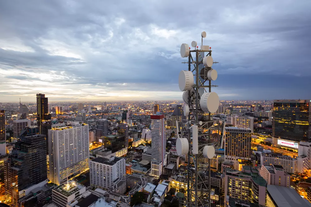 Foto de um horizonte urbano futurista ao entardecer, destacando uma torre de transmissão com diversos painéis de antenas, em uma cidade moderna.

