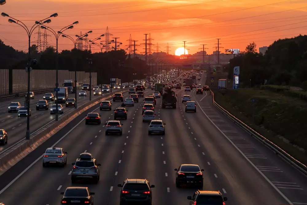 Tráfego intenso em uma rodovia ao pôr do sol, com vários carros e iluminação pública ao longo da estrada, criando uma cena urbana ao entardecer.
