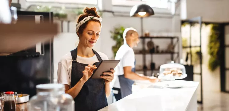Mulher sorridente, proprietária de cafeteria ou restaurante, usando avental e lendo um tablet, ao fundo uma colega trabalhando na cozinha, ambiente acolhedor e moderno.
