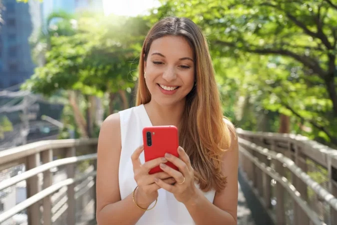 Mulher sorridente usando celular vermelho em ambiente ao ar livre, com árvores verdes ao fundo, transmitindo alegria e conexão digital.