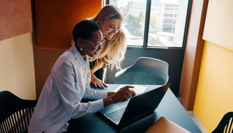 Duas mulheres de diferentes origens culturais colaborando em um projeto em uma sala com grande janela, promovendo diversidade que gera valor no ambiente de trabalho.