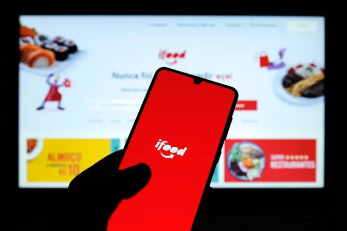 Pessoa segurando smartphone exibindo o aplicativo iFood, com tela mostrando o logo do serviço de delivery de comida, em frente a um computador com site do iFood aberto.