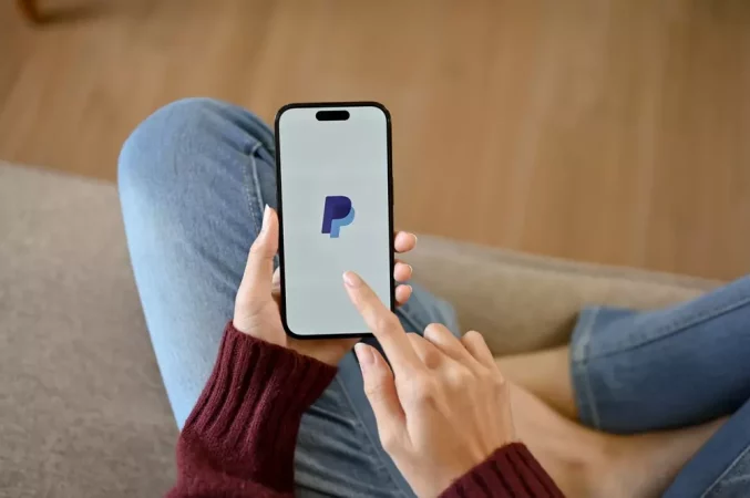 Pessoa usando smartphone com o logo do PayPal na tela, sentado em um sofá confortável, representando transações financeiras online com PayPal.