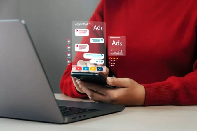 Pessoa segurando smartphone com visualizações de anúncios digitais e gráficos em um ambiente de trabalho com laptop ao fundo.